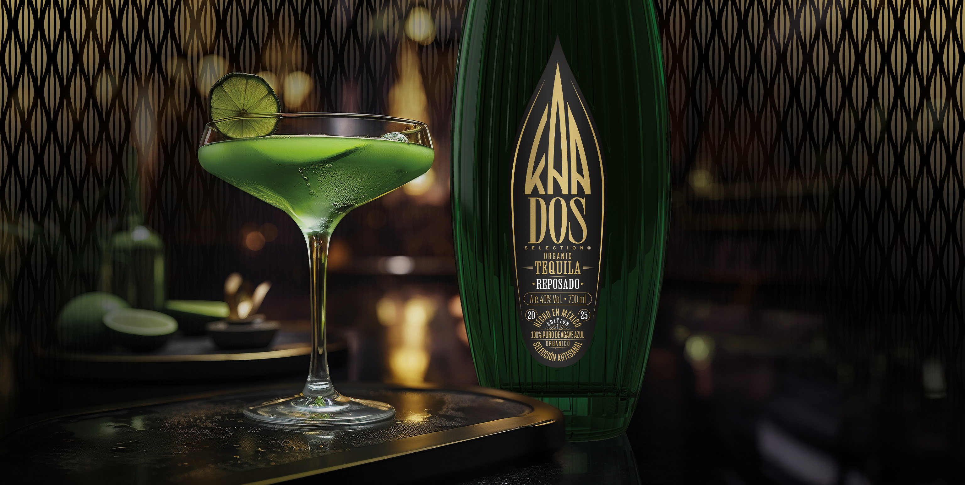 A close-up of a cocktail scene in a dimly lit bar setting. On the left, a green cocktail is served in a coupe glass, garnished with a fresh lime wheel on the rim. The drink sits on a black tray with a gold edge, which has condensation droplets. Behind it, sliced limes and bar tools are subtly visible. On the right, a large green glass bottle of "KAA DOS Tequila Reposado" dominates the frame, with a gold label reading "Organic Tequila Reposado – 100% Puro de Agave Azul – Selección Artesanal." The background