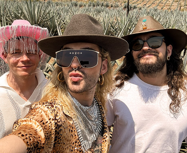 Three people taking a selfie in a sunny agave field. In the foreground, a person with long blond hair wears a brown hat, oversized sunglasses with gold accents, and a leopard-print shirt with a rhinestone scarf. On the left, someone smiles in a white top and a striking pink cowboy hat with silver fringe. On the right, a person with long dark hair, a beard, and earrings wears a white T-shirt, dark sunglasses, and a brown hat decorated with colorful pins. Rows of agave plants stretch into the background under