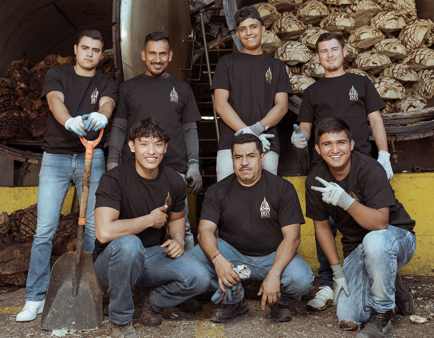 Group photo of seven male distillery workers posing in front of two large agave ovens, one filled with raw agave hearts and the other with roasted ones. All wear black "KUNA DOS" T-shirts, jeans, and work gloves. Two men kneel in front, one smiling with a peace sign. The others stand behind them, one leaning on a shovel. The mood is cheerful and proud, set in an industrial outdoor setting with visible agave remnants on the ground.