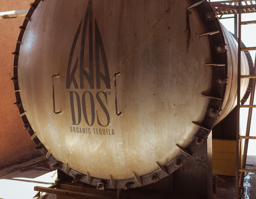 Close-up of the round metal door of an industrial agave oven at a tequila distillery. The door is tightly bolted and prominently features the black "KUNA DOS ORGANIC TEQUILA" logo printed vertically across the center. The oven's cylindrical body extends backward and is partially enclosed by a yellow concrete base and a metal scaffold. Warm lighting emphasizes the texture of the brushed metal surface.