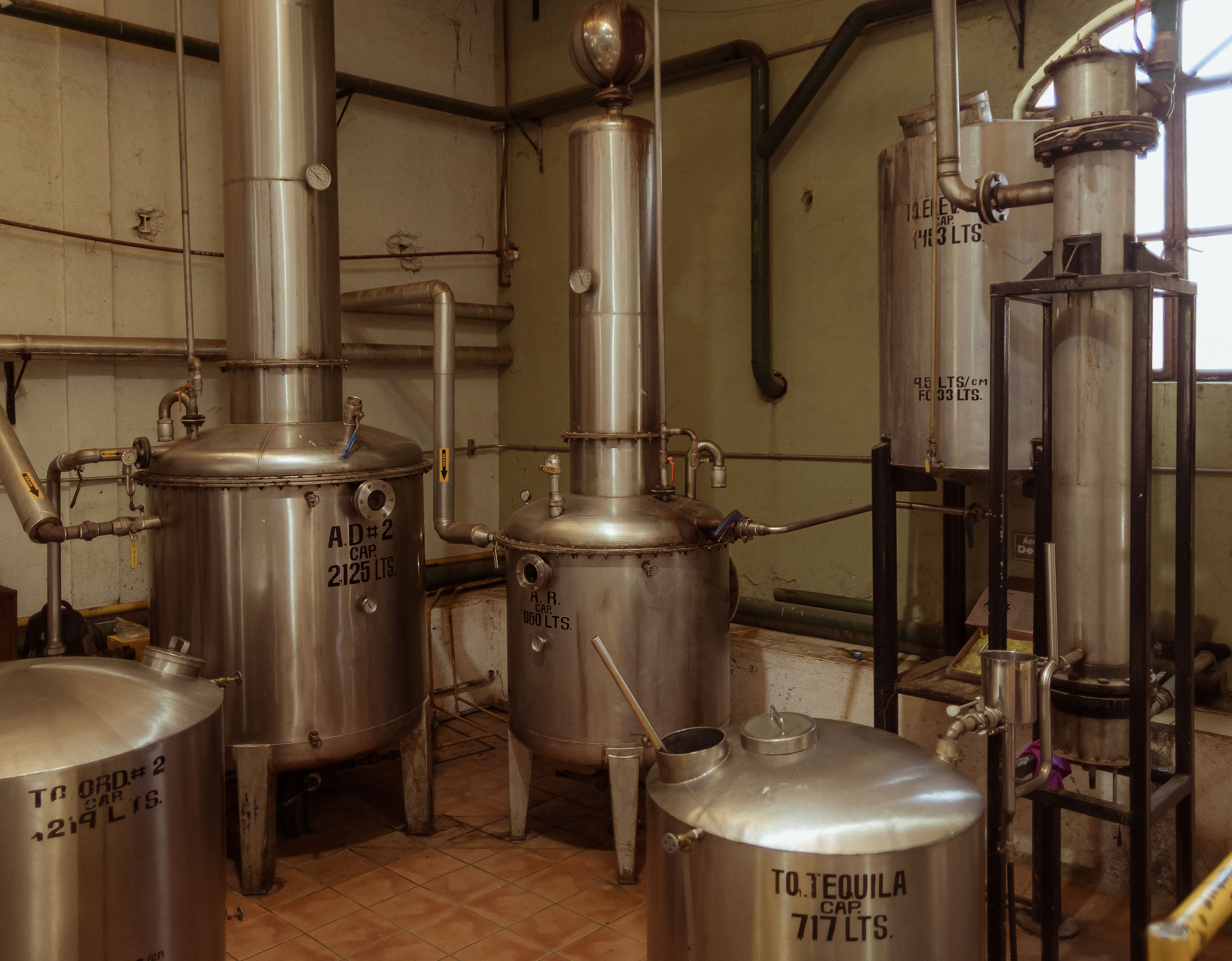 Industrial distillery setup with multiple stainless steel tanks and columns connected by metal pipes. The equipment is marked with black stencil lettering indicating capacities such as “2125 LTS”, “717 LTS” and “160 LTS”. The scene is well-lit with warm tones, showcasing the production environment for tequila. The machinery is placed on a tiled floor with green-painted walls in the background.
