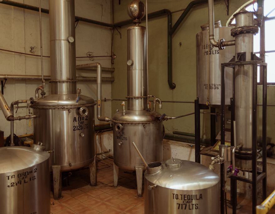 Industrial distillery setup with multiple stainless steel tanks and columns connected by metal pipes. The equipment is marked with black stencil lettering indicating capacities such as “2125 LTS”, “717 LTS” and “160 LTS”. The scene is well-lit with warm tones, showcasing the production environment for tequila. The machinery is placed on a tiled floor with green-painted walls in the background.