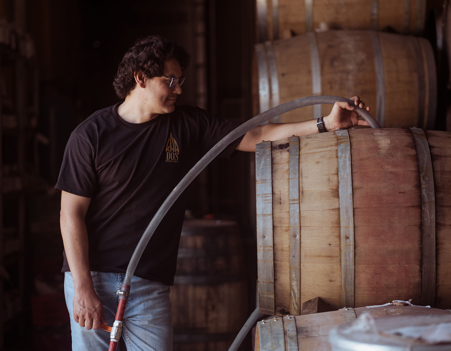 Man wearing a black KA DOS Organic Tequila T-shirt and jeans is filling a large wooden barrel with liquid using a thick hose. He is standing in a dimly lit distillery or warehouse space, gently resting one hand on the barrel and holding the hose with the other. The aged wooden barrels with metal bands and the subdued lighting convey a sense of traditional craftsmanship in tequila production.