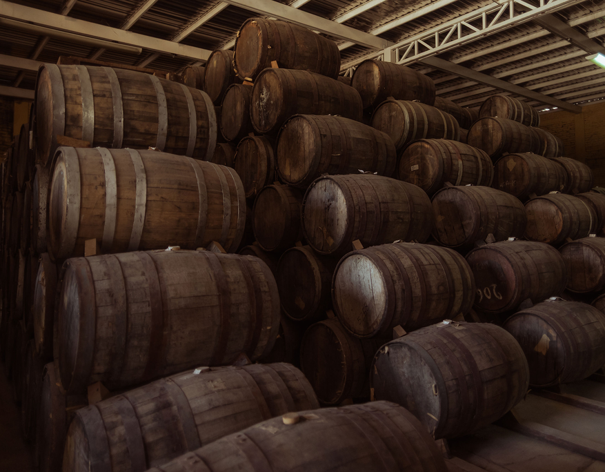 Interior view of a tequila aging warehouse with dozens of stacked wooden barrels. The barrels are arranged in rows on wooden blocks, showing signs of age and use. The industrial ceiling structure above suggests a controlled environment for aging. The lighting is warm and subdued, emphasizing the traditional and artisanal atmosphere of the space.