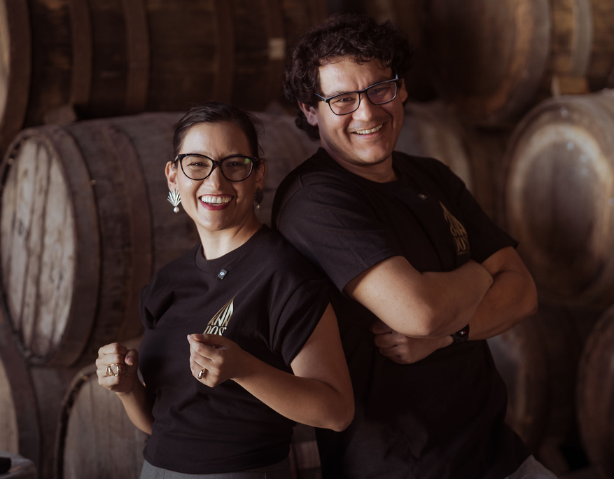 Two smiling individuals stand back-to-back in front of stacked wooden tequila barrels. Both wear black T-shirts with the KNA DOS logo. The woman on the left wears agave-shaped earrings and red lipstick, clenching her fists in a joyful, celebratory gesture. The man on the right has curly hair and glasses, with arms crossed and a wide grin. The setting has a warm, rustic feel, highlighting the artisanal tequila environment.