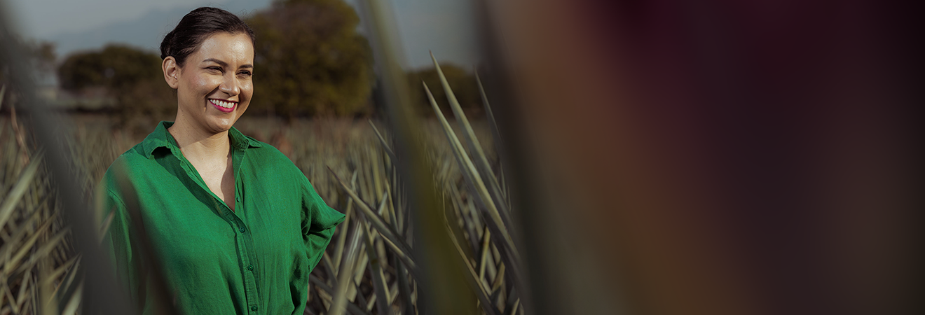Portrait of a smiling woman standing in an agave field. She wears a vibrant green shirt and has her dark hair tied back. Agave plants frame the image in the foreground and stretch into the distance. A soft, warm light enhances the natural tones of the scene, and blurred elements on the right side suggest a vignette or gradient overlay. The mood is open and welcoming.