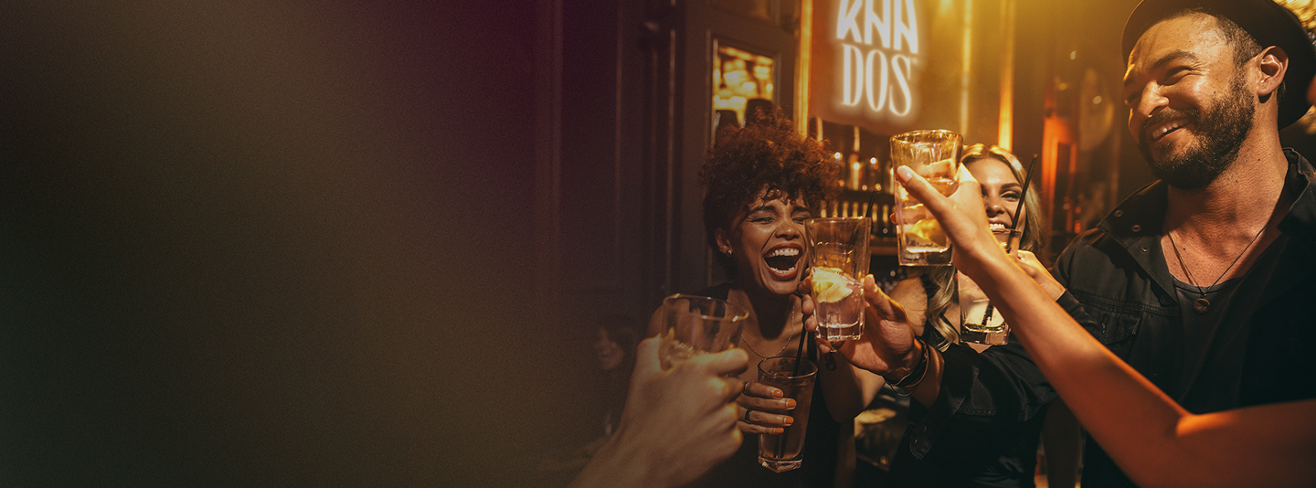 A group of friends is toasting with tall glasses at a lively bar. Everyone is smiling and laughing, enjoying the moment. The lighting is warm and golden, giving the scene a cozy and celebratory atmosphere. In the background, a softly glowing sign with the logo "KAA DOS" can be seen, adding a branded touch to the setting. The image captures the spirit of fun, friendship, and premium tequila enjoyment.